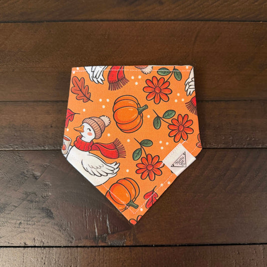 Orange pet bandana with white geese holding coffees and wearing knit hats and scarves with pumpkins and flowers in the background. 