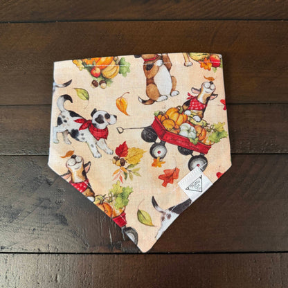 Cream pet bandana with dogs, autumn leaves, and a red wagon with a dog and pumpkins in it.