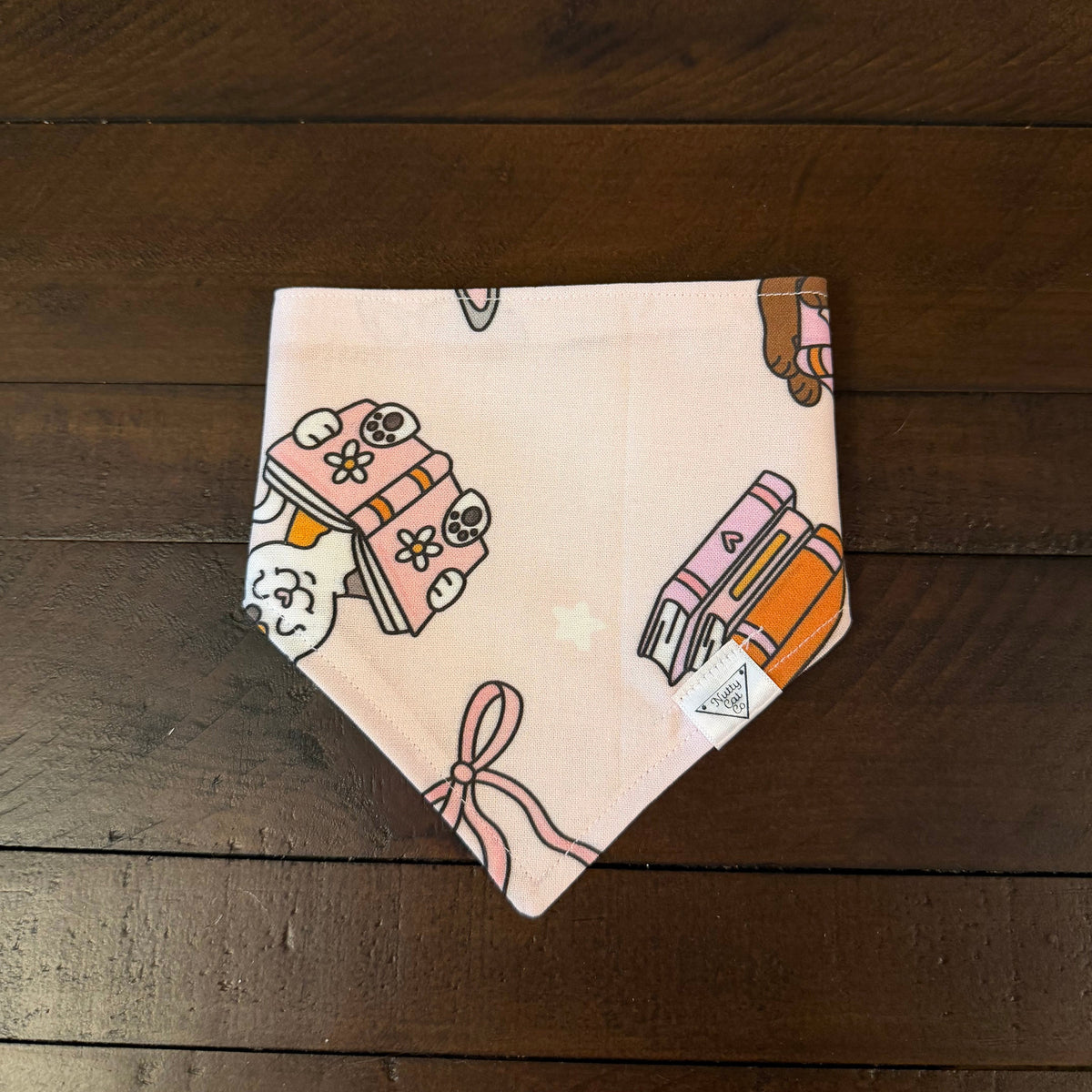 Pale pink pet bandana with a cat peeking over books, book stacks, and a ribbon bow.