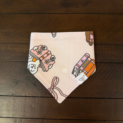 Pale pink pet bandana with a cat peeking over books, book stacks, and a ribbon bow.