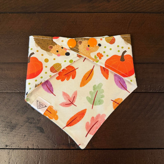 Cream pet bandana with hedgehogs, orange pumpkins, acorns, and colorful dots.